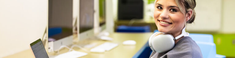 a smiling student with headphones around her neck sits at a laptop